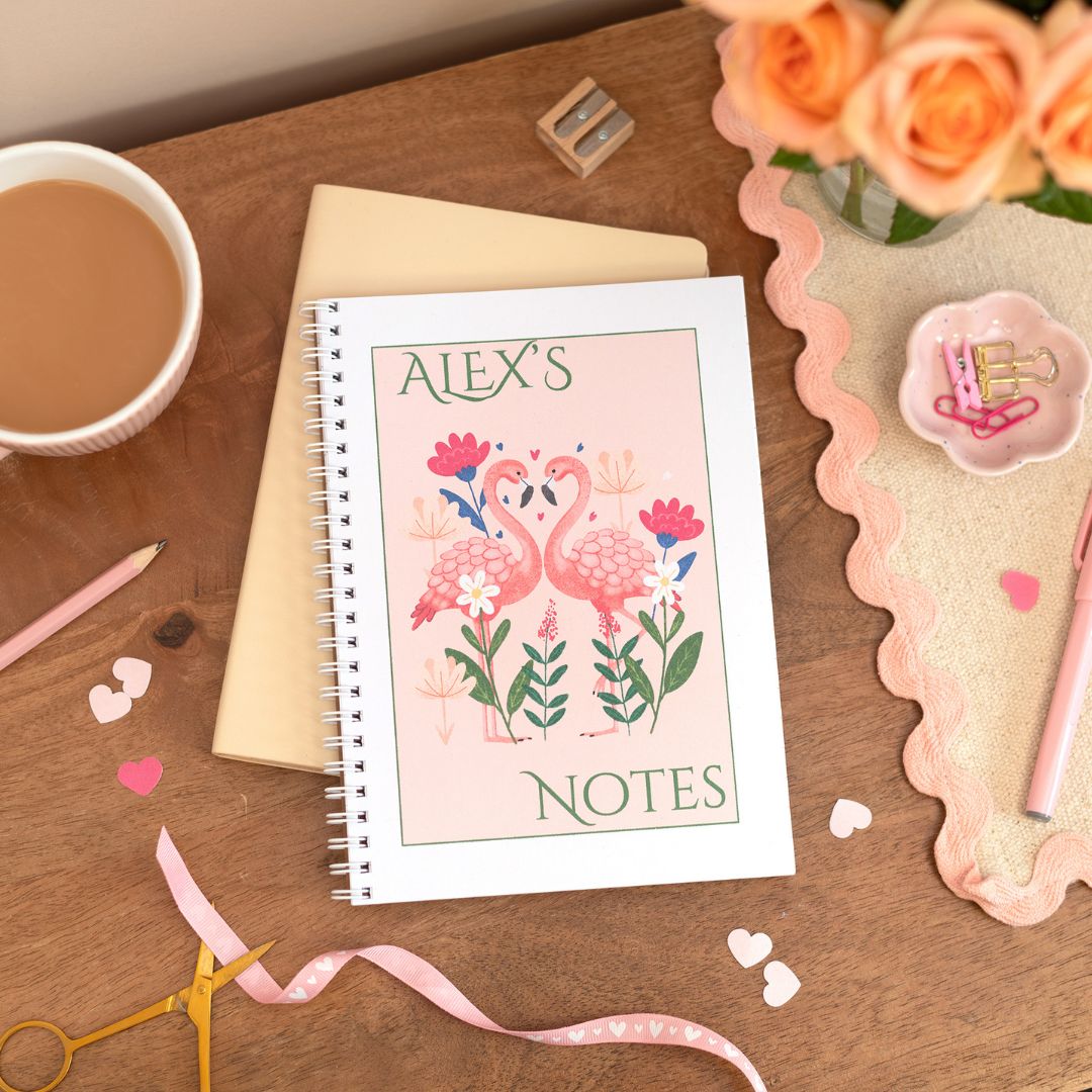 Personalised A5 notebook with flamingo design on the cover, placed on a desk next to a cup of coffee, pens, and decorative ribbons.