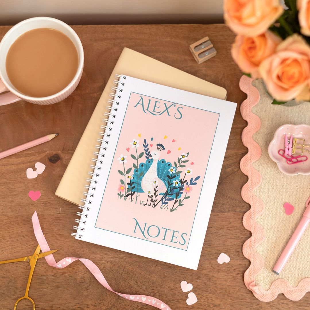Personalised A5 notebook with a peacock design on the cover, placed on a desk next to a cup of coffee, a pen, and some decorative ribbons.