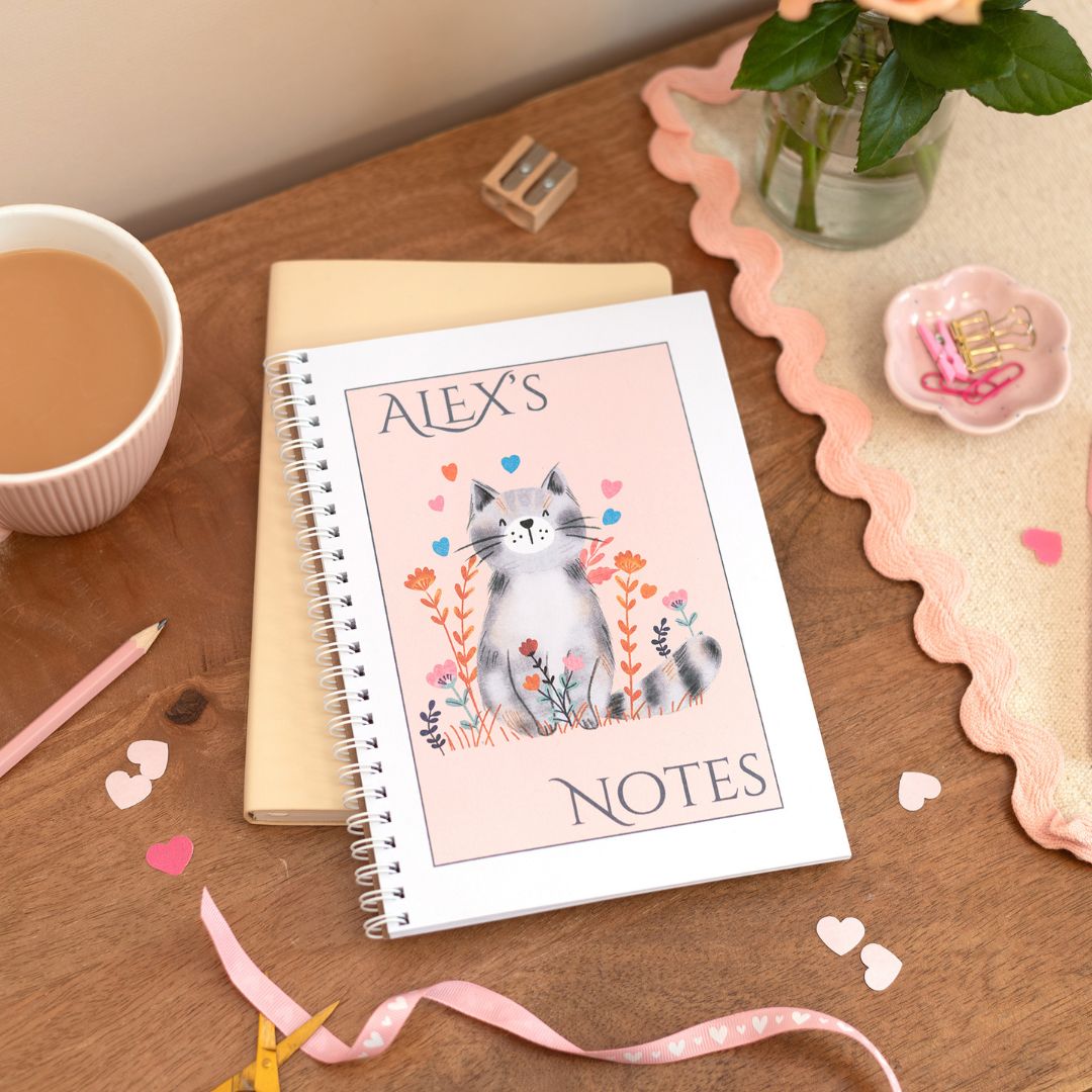 Personalised A5 notebook with a cute cat design and floral elements, set against a pink background, placed on a desk next to a cup of coffee and other stationery items.