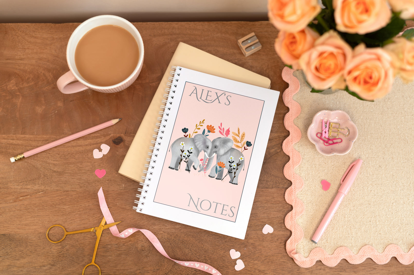 Personalised A5 Notebook which is wire bound. Features two elephants on the front against a pink background. The notebook is on a wooden table with a cup of coffee, flowers, and stationery items.