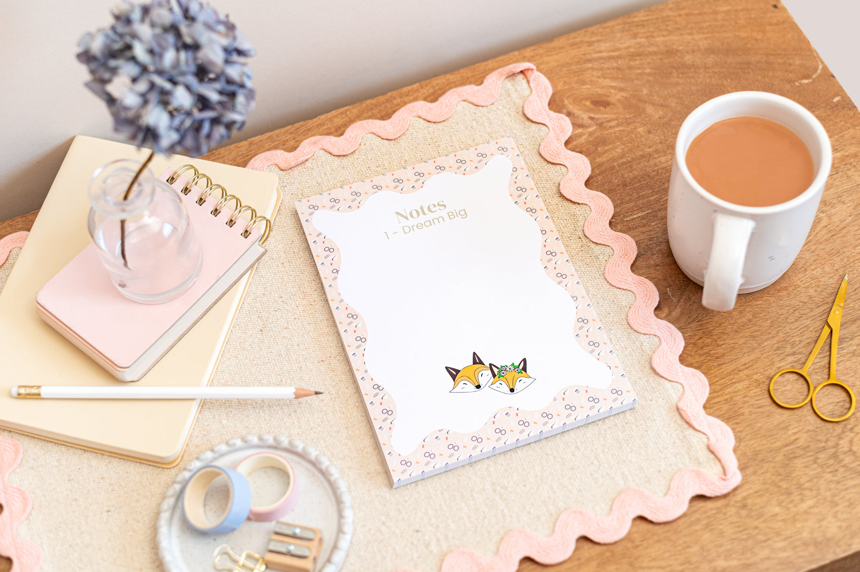An A5 daily planner pad with a floral and fox design on the cover, placed on a wooden desk next to a cup of tea, a notebook, pens, and scissors.