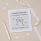 A congratulations card for a grandson's engagement with two celebratory glasses on the front, placed on a neutral background with a white ribbon, pen, and scattered confetti. The text on the card reads "congratulations to our grandson and wife to be on your engagement"