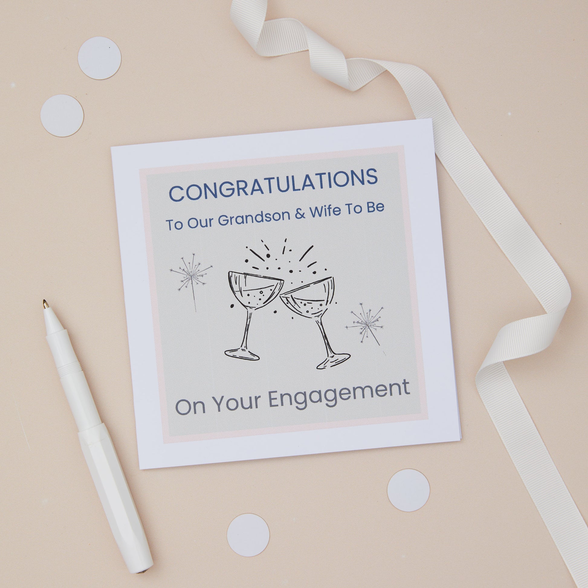 A congratulations card for a grandson's engagement with two celebratory glasses on the front, placed on a neutral background with a white ribbon, pen, and scattered confetti. The text on the card reads "congratulations to our grandson and wife to be on your engagement"