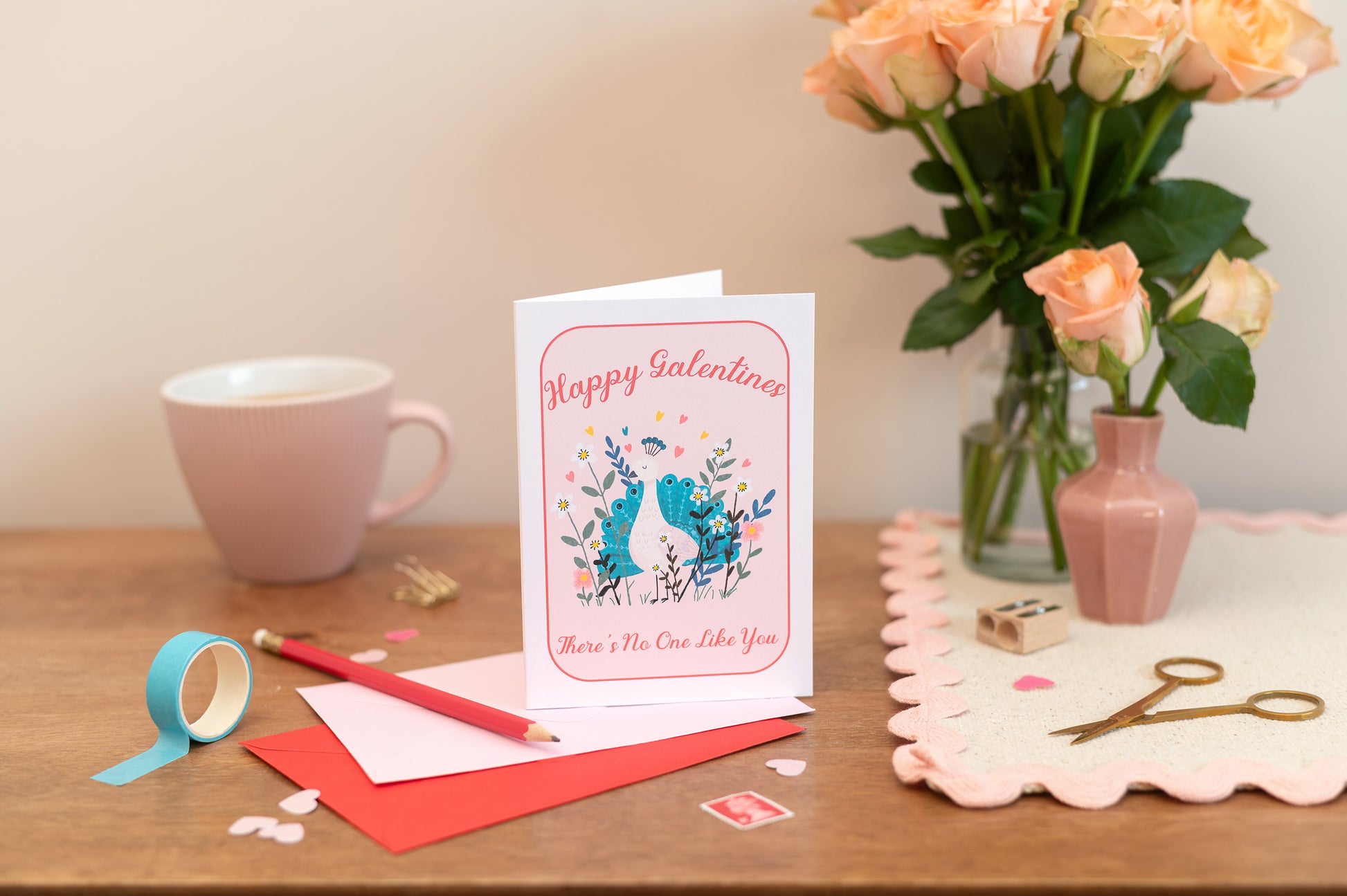 A greeting card with 'Happy Galentine's Day' text and a whimsical peacock illustration , placed on a table with a floral arrangement, a mug, and decorative items.