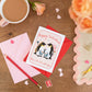 Happy Valentines Penguins Romance Card with pink and red envelopes. The card sits on a desk with stationery and a cup of tea. There is floral roses on the desk