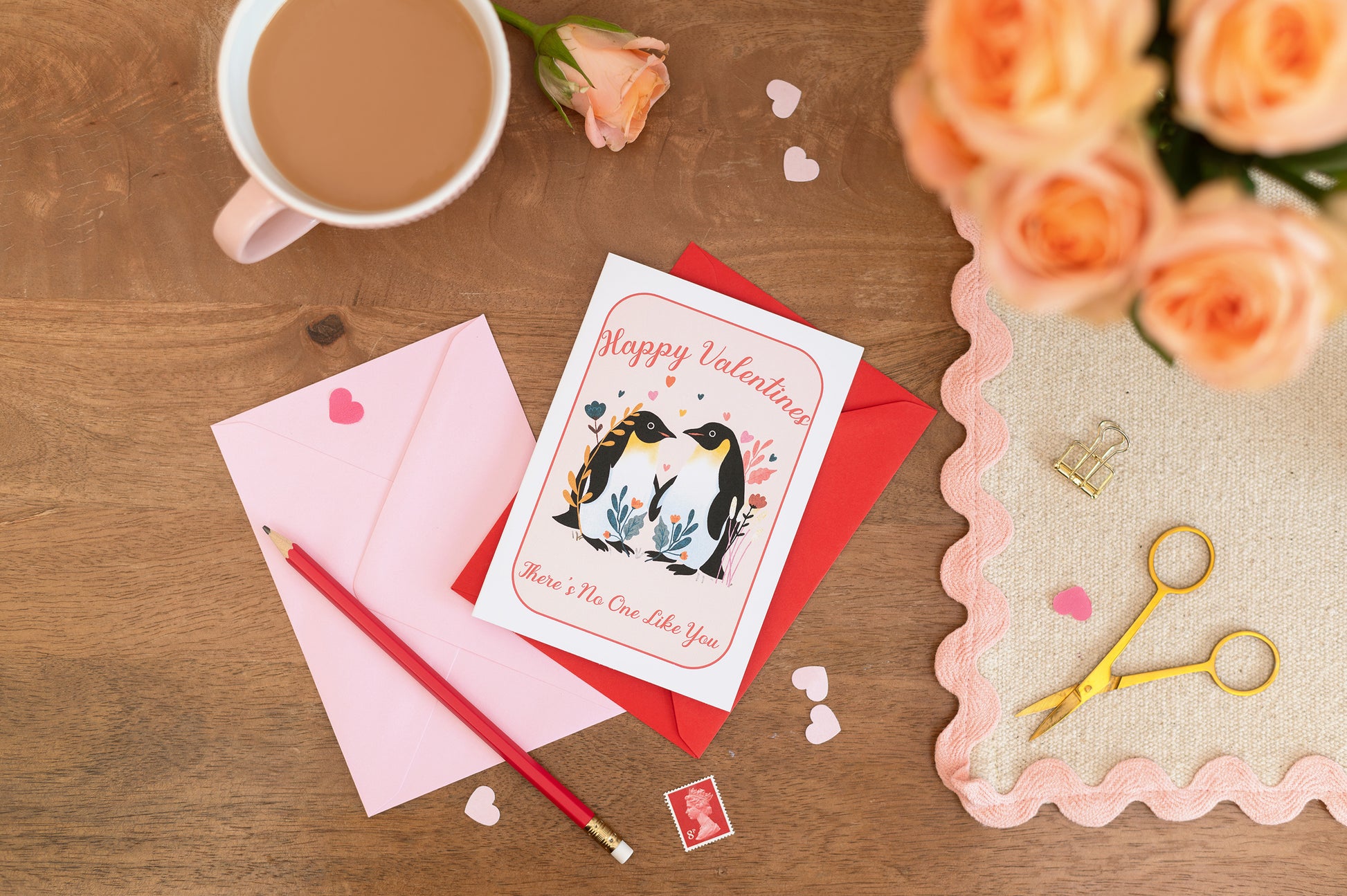 Happy Valentines Penguins Romance Card with pink and red envelopes. The card sits on a desk with stationery and a cup of tea. There is floral roses on the desk