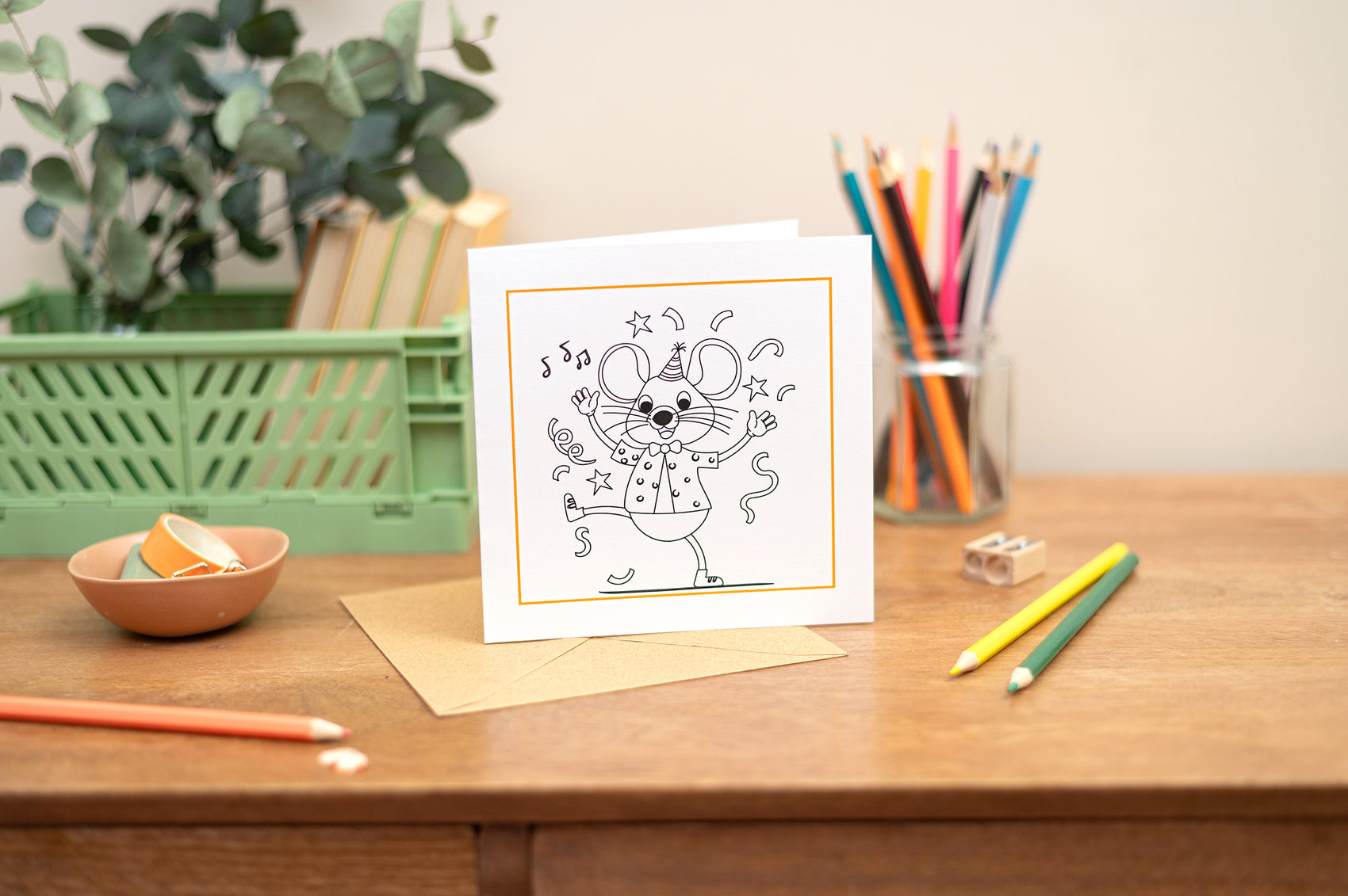 Kids Colouring in Card featuring a party mouse with music notes and party elements around it. The Card is placed on a wooden table with stationery elements surrounding it.