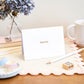 A folded 'Thank You' letterpress card on a wooden table with a cup of tea, a flower vase, and other stationery items in the background.