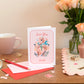 A Valentine's Day Romance and Friendship Card with 'Love You Loads' text with a cute bear holding a bunch of flowers. The Card is placed on a table alongside a pink cup, with a vase of pink roses and decorative items.