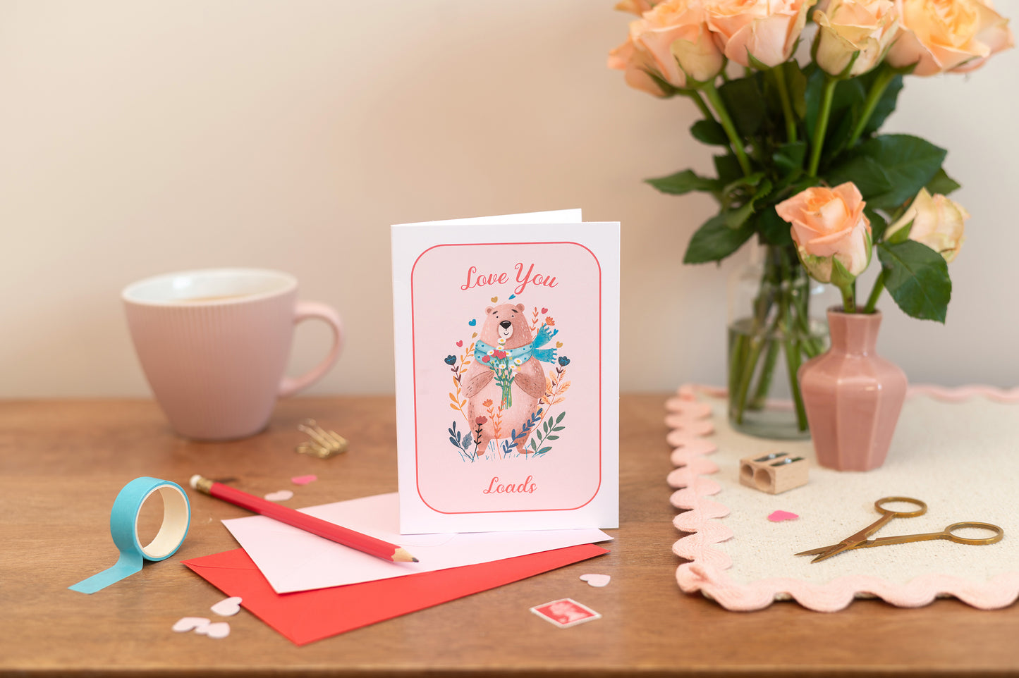 A Valentine's Day Romance and Friendship Card with 'Love You Loads' text with a cute bear holding a bunch of flowers. The Card is placed on a table alongside a pink cup, with a vase of pink roses and decorative items.