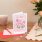 A Valentines Romance Friendshop greeting card with a flamingo design, along with an envelope and a pair of scissors, placed on a table with a vase of pink roses in the background.  The text says "we make the perfect pair"