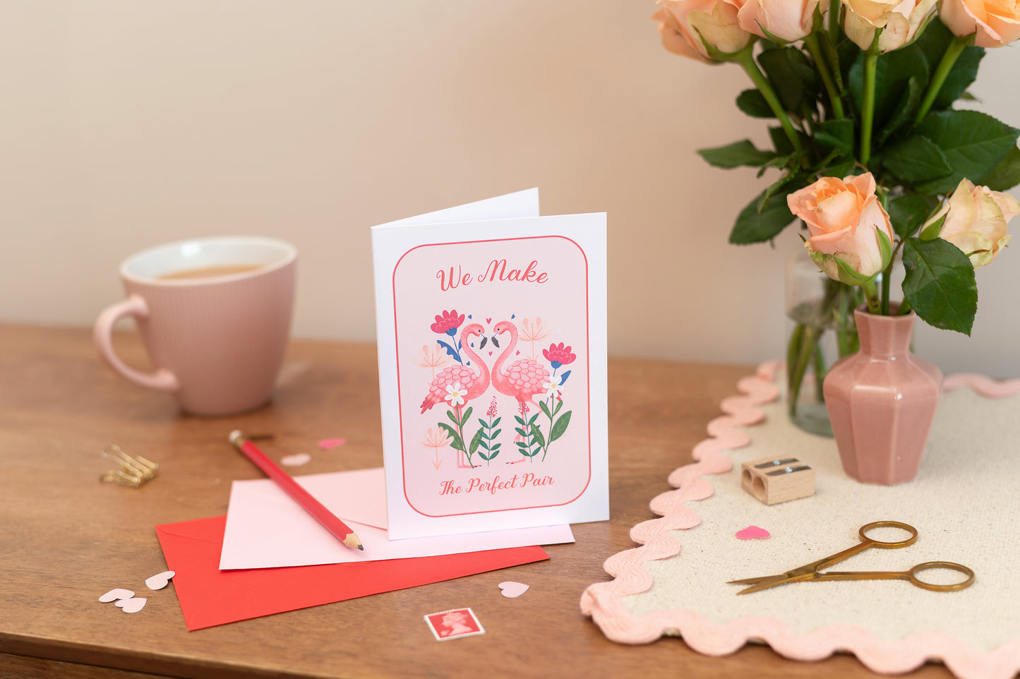 A Valentines Romance Friendshop greeting card with a flamingo design, along with an envelope and a pair of scissors, placed on a table with a vase of pink roses in the background.  The text says "we make the perfect pair"