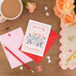 A Valentine's Day themed greeting card with an illustration of two elephants facing each other with the text "you're my favourite person", placed on a table alongside a mug of tea, a red rose, and a pair of scissors.