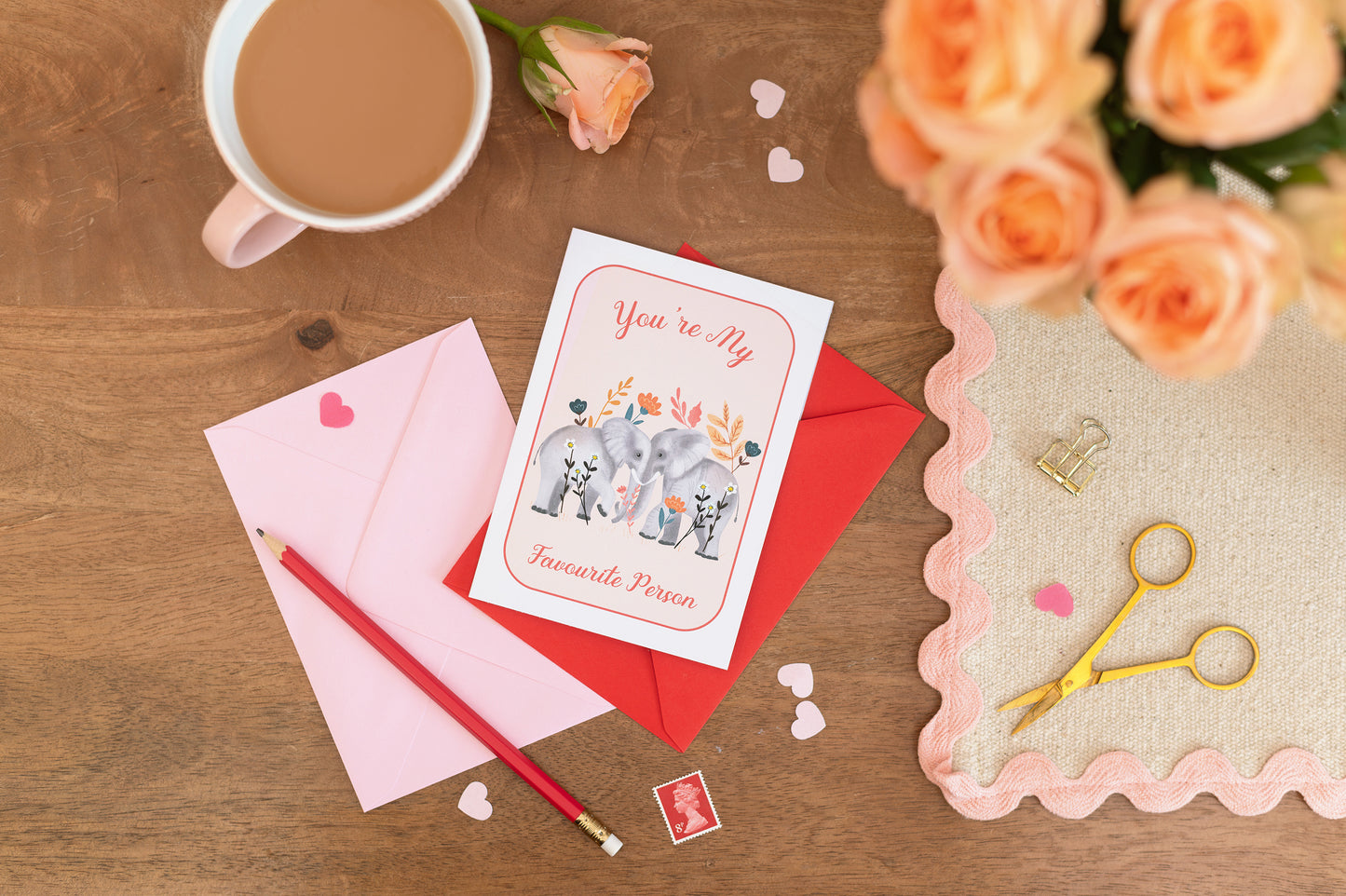 A Valentine's Day themed greeting card with an illustration of two elephants facing each other with the text "you're my favourite person", placed on a table alongside a mug of tea, a red rose, and a pair of scissors.