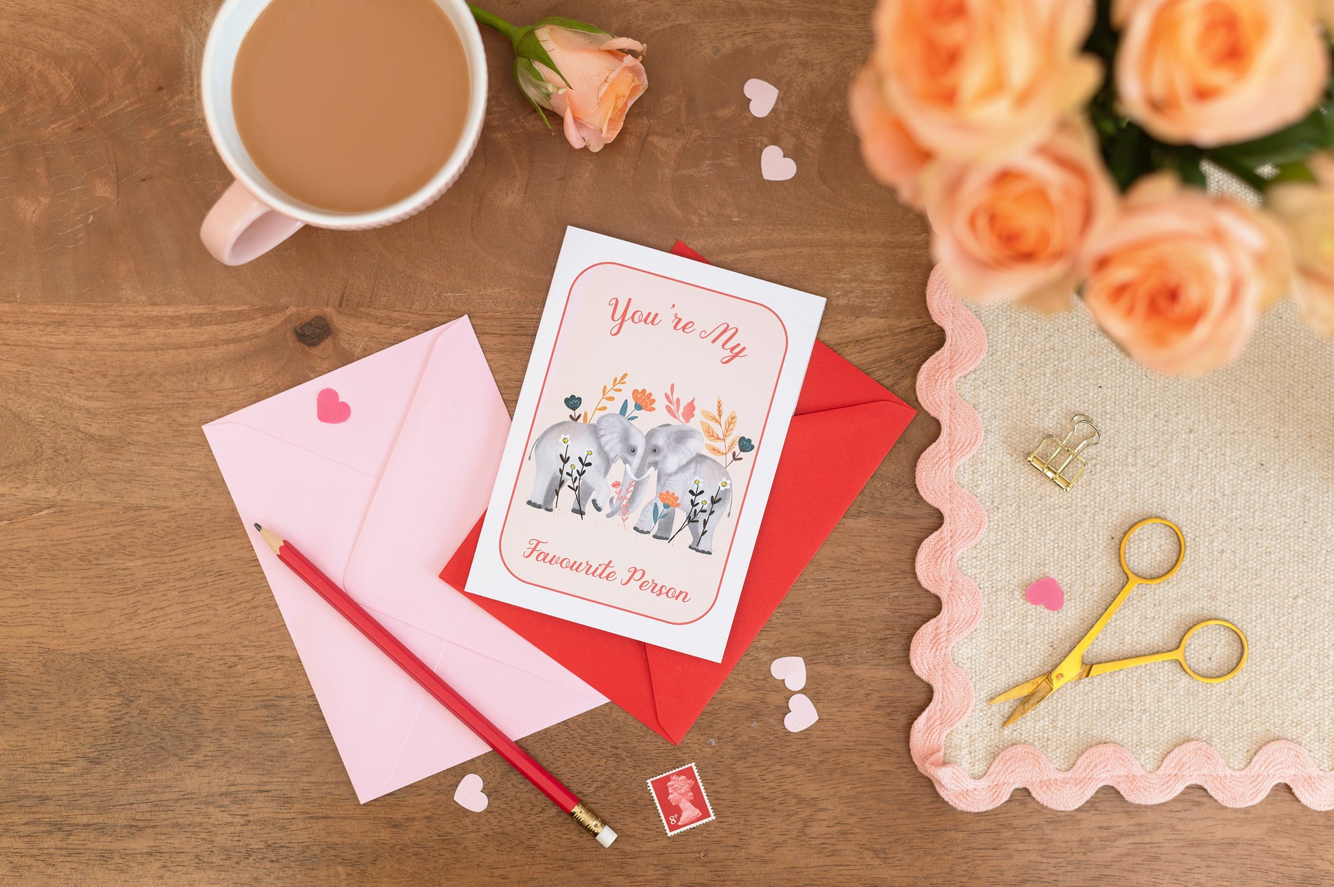 A Valentine's Day themed greeting card with an illustration of two elephants facing each other with the text "you're my favourite person", placed on a table alongside a mug of tea, a red rose, and a pair of scissors.