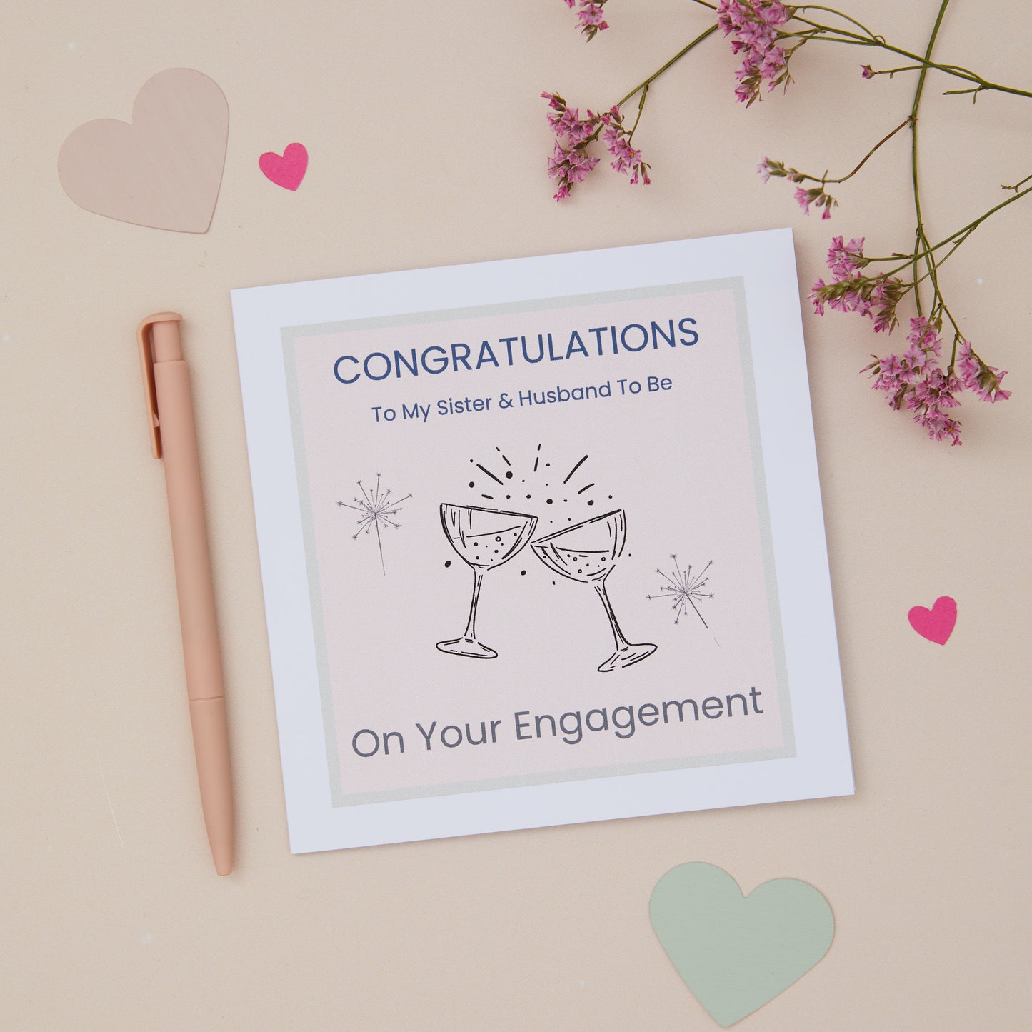 A congratulations card for a sister's engagement featuring two celebratory glasses, with a pink pen, and decorative flowers on a pastel background.