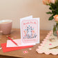 A greeting card with a cartoon image of a cat and the text 'THANKS FOR BEING SUCH A GREAT FRIEND' on it, accompanied by a vibrant pink envelope and a pair of scissors for opening the envelope, placed on a table with a floral decoration in the background.