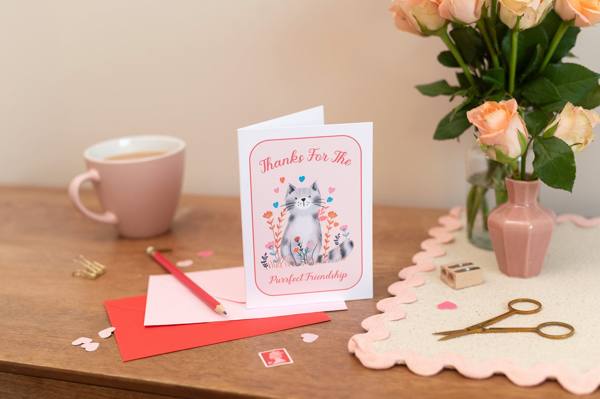 A greeting card with a cartoon image of a cat and the text 'THANKS FOR BEING SUCH A GREAT FRIEND' on it, accompanied by a vibrant pink envelope and a pair of scissors for opening the envelope, placed on a table with a floral decoration in the background.